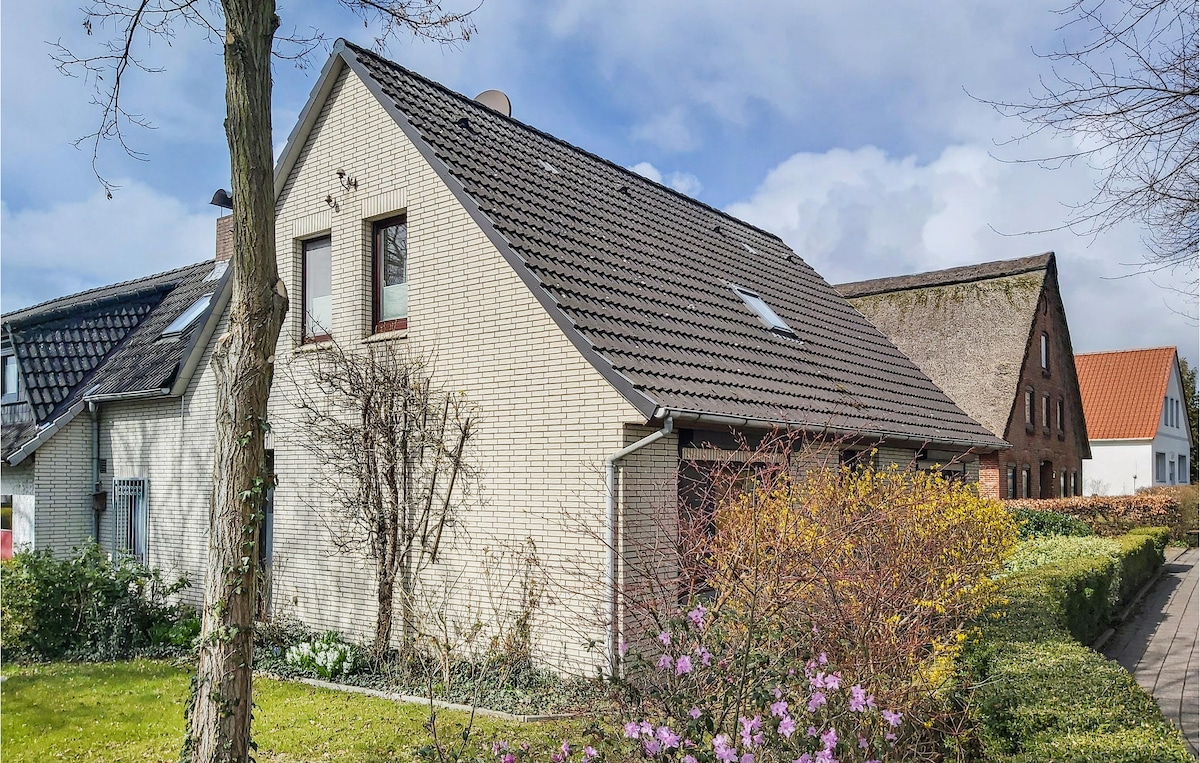 The exterior of the cottage is shown, featuring a modern brick facade with multiple windows. Greenery surrounds the home, including blooming flowers and manicured hedges. The well-maintained yard offers a welcoming entrance to this vacation home.