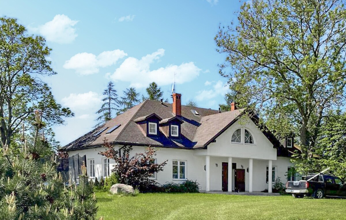 A spacious holiday home is set against a clear blue sky, surrounded by lush greenery. The building features a large arched entrance and multiple gabled roofs, complemented by a well-kept lawn and mature trees. A car is parked nearby, emphasizing the property's accessibility.