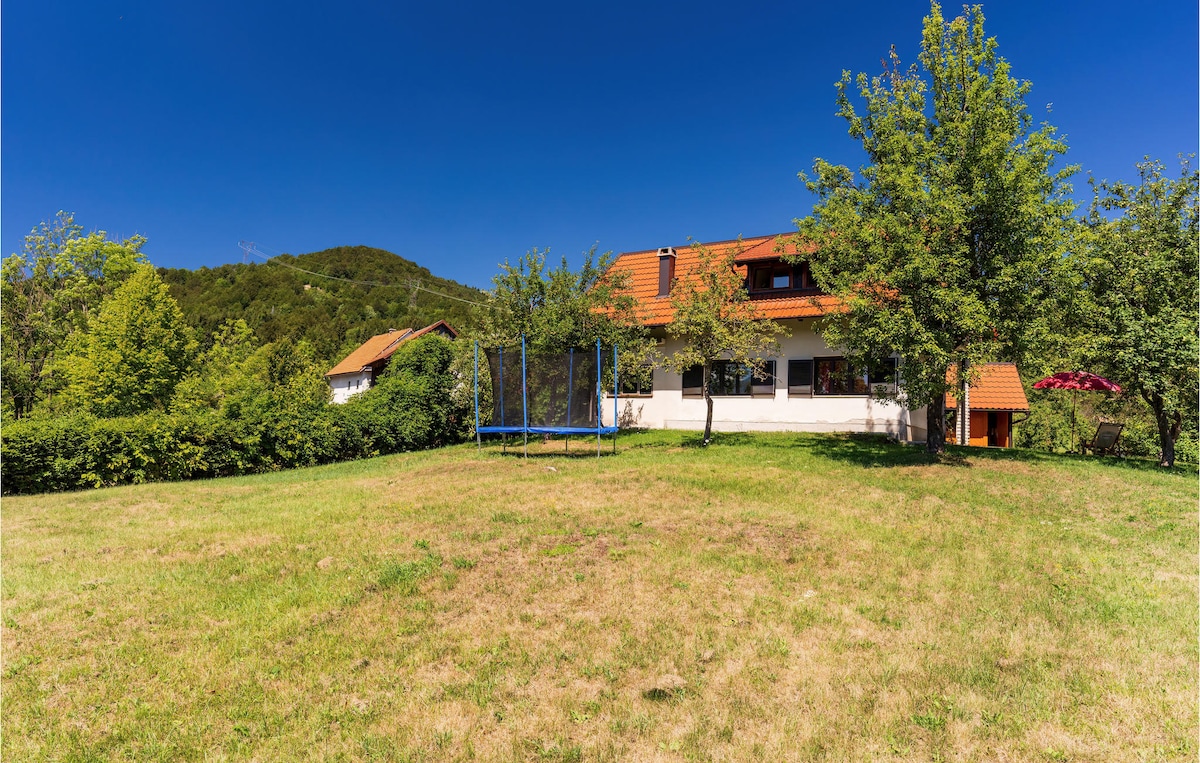 A spacious yard surrounds a charming holiday home, with lush greenery and trees providing a natural setting. A trampoline is visible in the yard, inviting outdoor play. The backdrop features rolling hills under a clear blue sky.