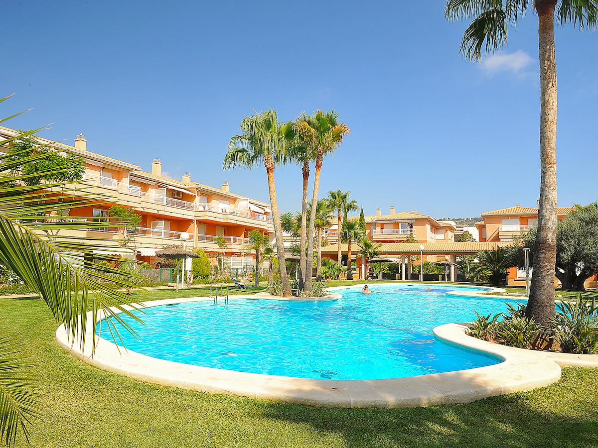 The kidney-shaped swimming pool is surrounded by lush palm trees and well-maintained lawns. Light blue water reflects the clear sky. Nearby buildings feature balconies and greenery, creating a serene environment for relaxation.