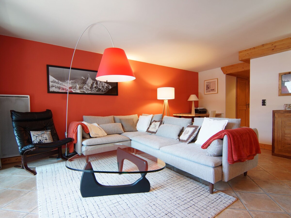 A spacious living room features a light gray sectional sofa adorned with decorative pillows, paired with a sleek black coffee table. A large red lampshade casts a warm light, complemented by a second floor lamp. The burnt orange wall enhances the cozy atmosphere of the space.
