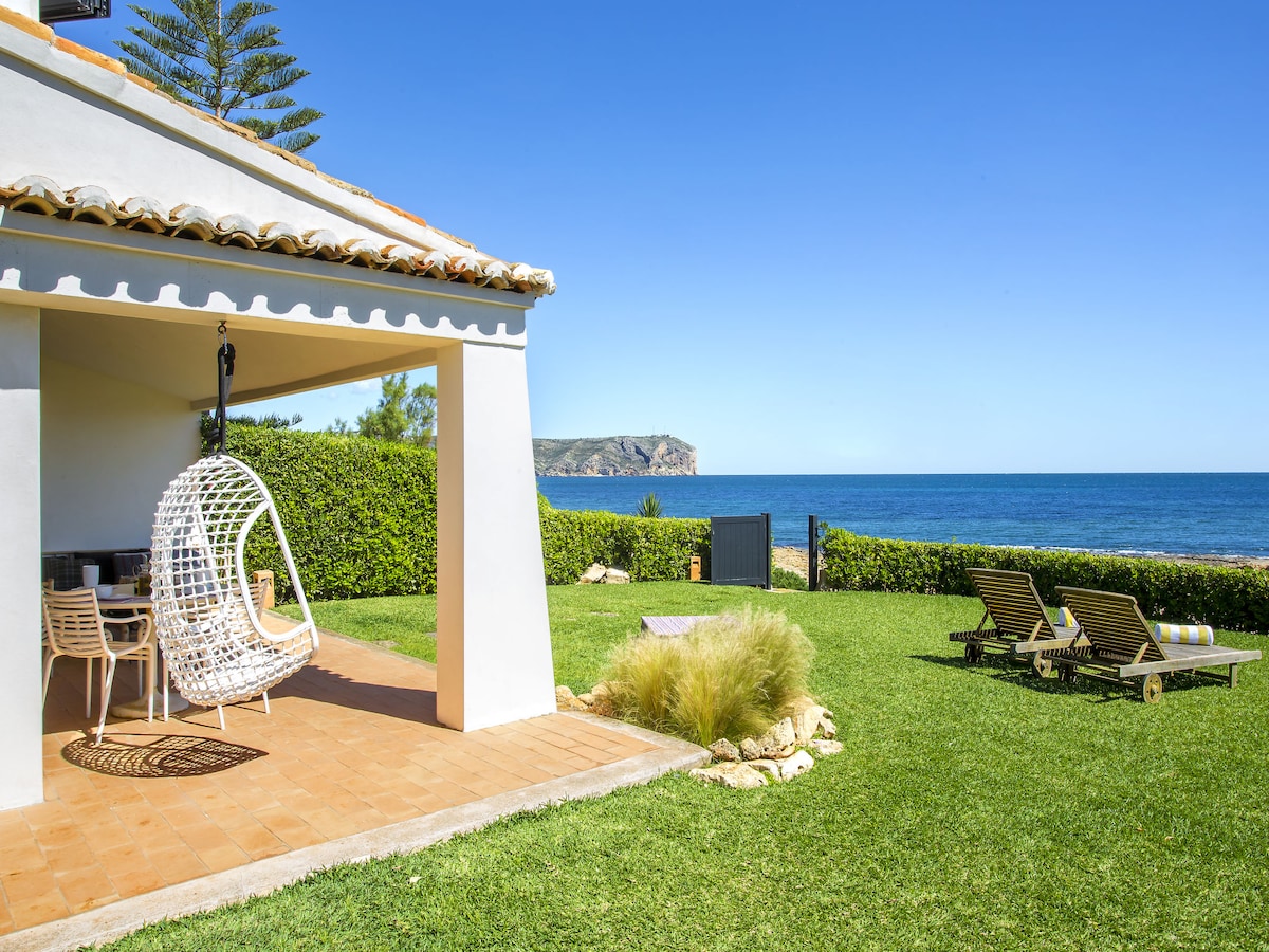 A patio area features a hanging chair and a dining set, overlooking a well-kept garden. Sun loungers provide relaxation options, while the expansive sea view stretches beyond. Lush greenery frames the scene, creating a serene outdoor space for enjoyment.