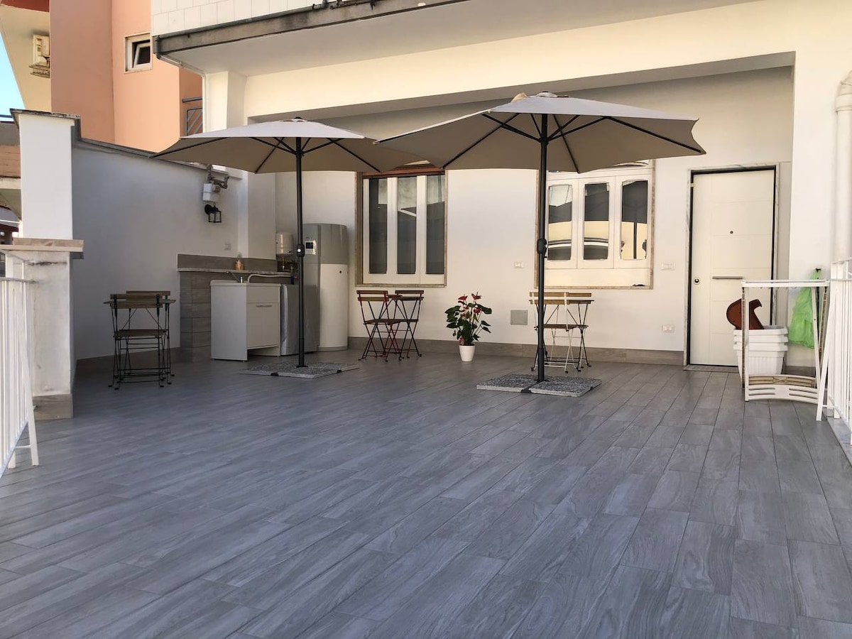 A spacious terrace is equipped with two large umbrellas providing shade, featuring a small dining area with two wooden chairs and a table. A barbecue grill is positioned nearby, and a door leads to the interior, complemented by a plant placed on the ground.