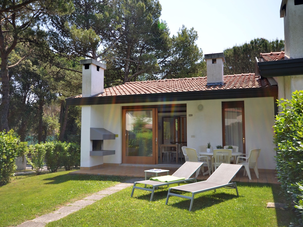 A welcoming terrace features comfortable loungers and a dining table surrounded by greenery. The entrance leads to large glass doors, showcasing a cozy interior. The area is bordered by well-maintained lawn and trees, creating a serene outdoor environment.