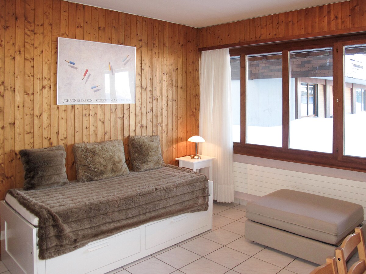 The living area features wooden walls and large windows allowing natural light to fill the space. A pull-out bed is positioned against the wall, accompanied by a cozy throw and decorative pillows. A lamp sits on a bedside table, complementing the inviting atmosphere.