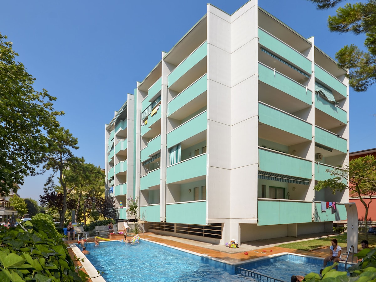 The exterior of a multi-story apartment building with a light turquoise façade is shown. A swimming pool is situated in the foreground, surrounded by greenery. Balconies with hanging laundry are visible, indicating the presence of several units within the residence.