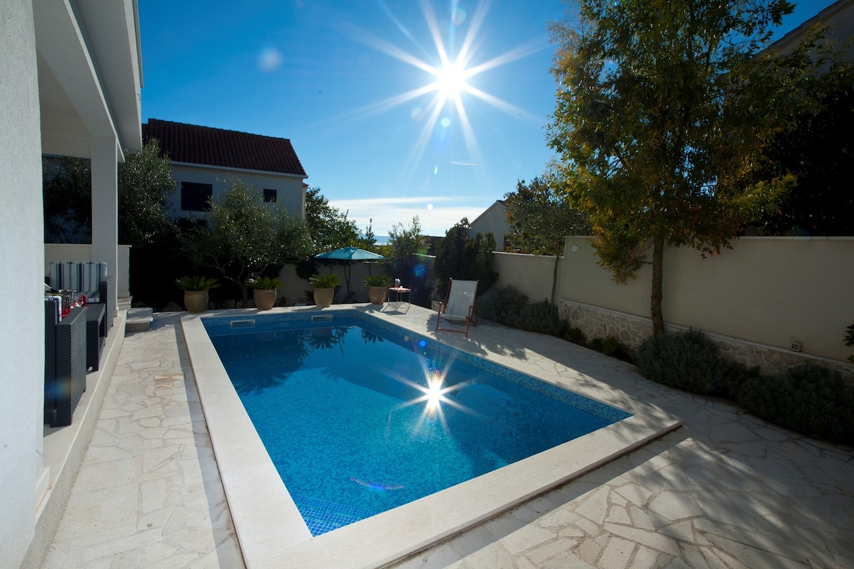 The outdoor area features a private swimming pool, reflecting sunlight with sparkling highlights. Surrounding the pool are stone tiles and well-maintained greenery, including potted plants. A loungers is positioned nearby, inviting relaxation in a serene environment.