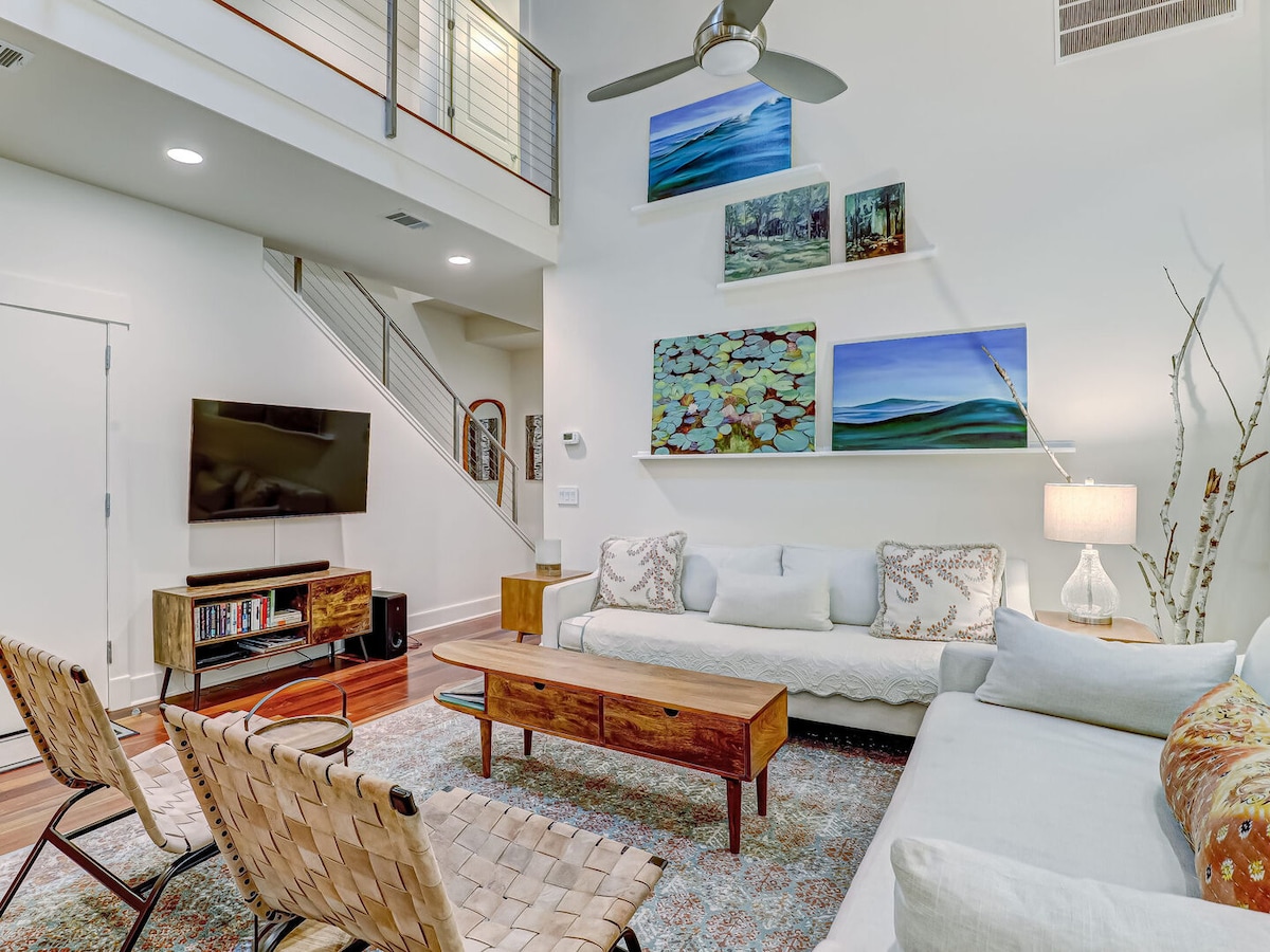 A bright living area is showcased with tall ceilings and large artwork adorning the walls. A spacious light-colored sofa and matching armchairs compliment the wood coffee table. Natural light floods in from the windows, creating a refreshing and open ambiance.