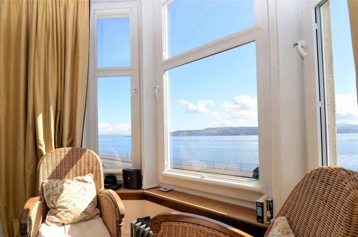 A bright corner of the living room features two wicker chairs positioned near large bay windows, offering panoramic views of the Clyde Estuary. Soft natural light fills the space, enhancing the tranquil atmosphere while the sea and sky create a captivating backdrop.