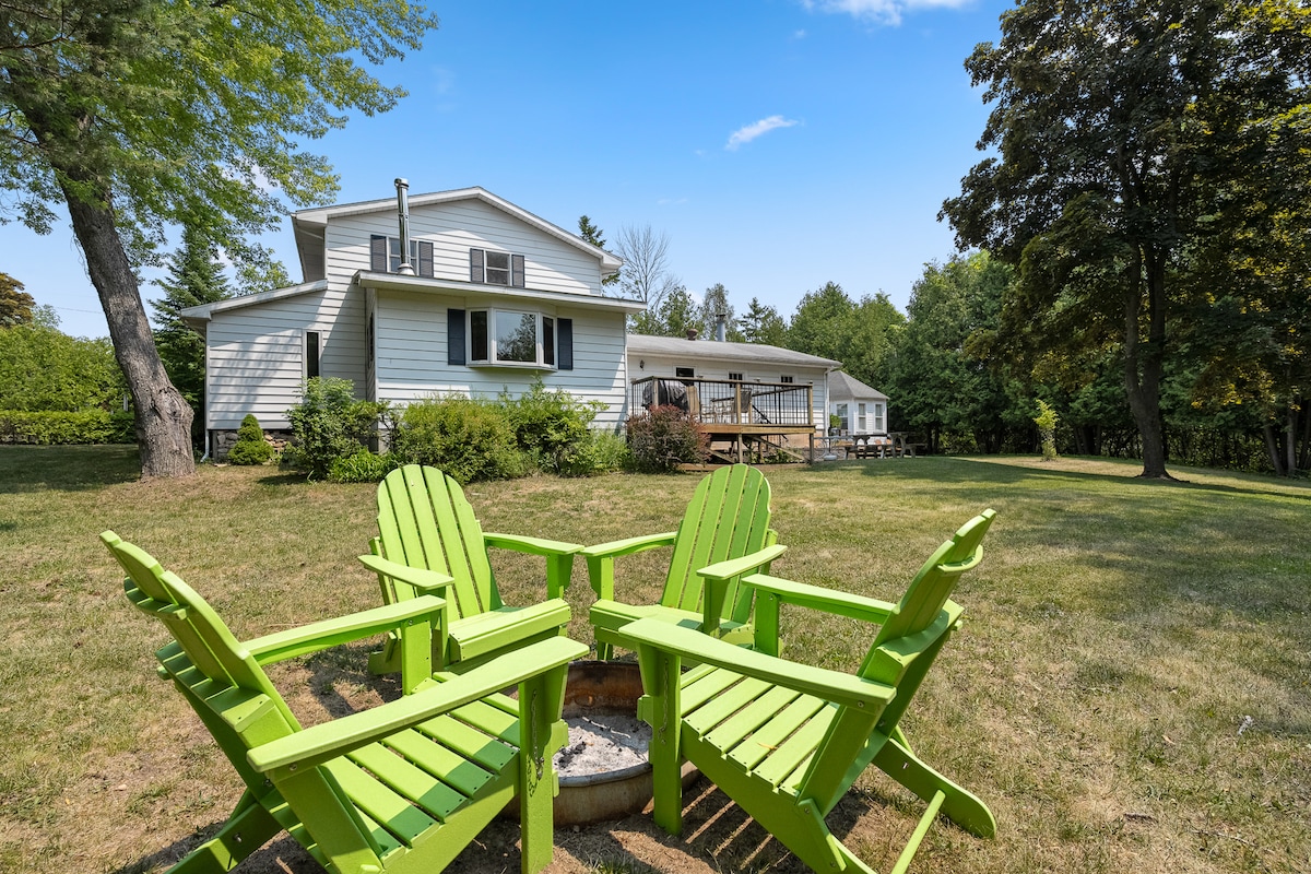 A spacious backyard is highlighted by four vibrant green adirondack chairs arranged around a fire pit. A large home is visible in the background, featuring a raised deck with outdoor seating and surrounded by trees and greenery.