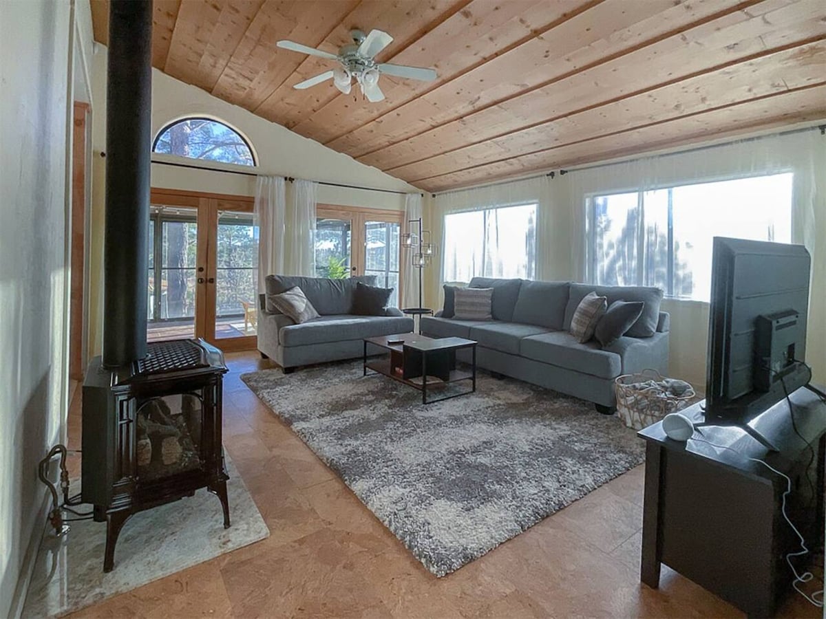 A spacious living area features a light-colored wood ceiling and large windows, allowing natural light to fill the room. Comfortable seating arrangements are provided by a sectional sofa and additional chairs. A compact table sits on a plush area rug, while a television and a heater are positioned nearby.