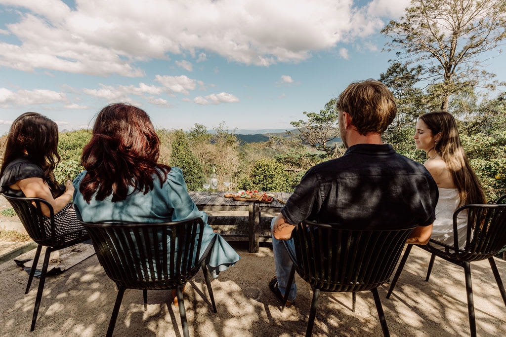 Four individuals sit in black outdoor chairs, facing a scenic view of the mountains and blue sky. A table with refreshments is visible in front of them. The surrounding landscape features lush greenery, suggesting a serene outdoor setting.