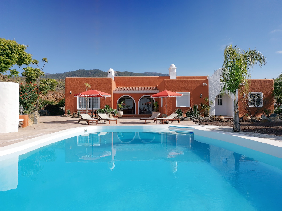 A spacious outdoor area features a sparkling private pool, reflecting the vibrant colors of the villa's exterior. Two red umbrellas provide shade over lounge chairs arranged on the sunlit patio, while lush landscaping and mountains can be seen in the background.
