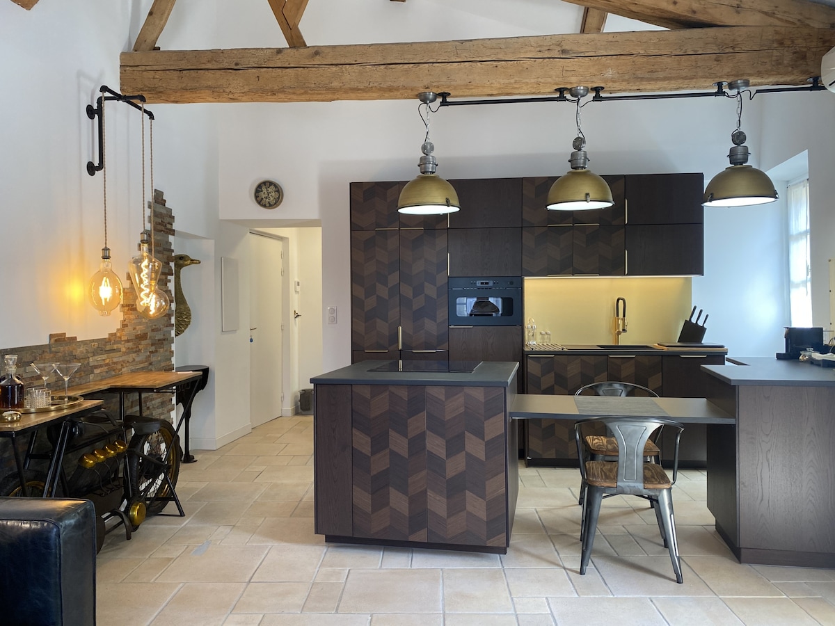 The kitchen features a modern design with wooden cabinetry and geometric patterns. High ceilings with exposed beams are complemented by industrial-style pendant lights. A large island with seating is present, alongside a range of appliances and dishware. The space is bright and airy, enhancing functionality.