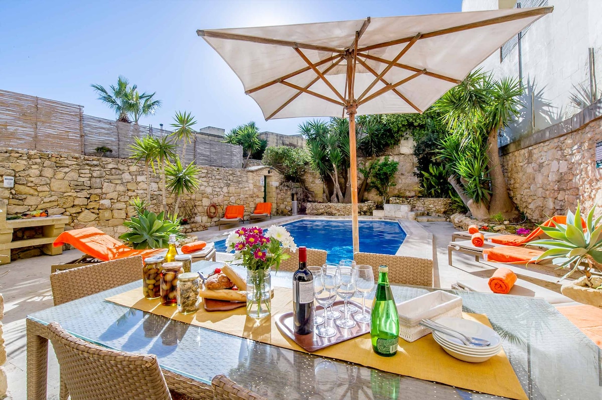 An outdoor dining area is set with a glass table, complete with plates, glasses, and a bottle of sparkling water. A large umbrella provides shade over the table. Vibrant orange loungers surround the private swimming pool, framed by palm trees and lush greenery.