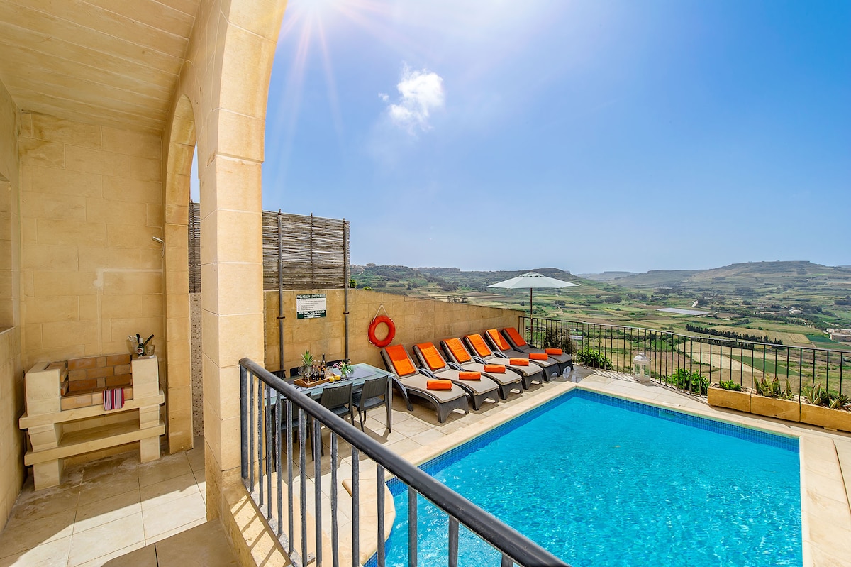 A private outdoor area features a clear blue pool surrounded by sun loungers with orange cushions. A shaded umbrella offers respite from the sun, while a scenic view of the countryside enhances the tranquil atmosphere, complemented by a stone wall and a small wooden bench.