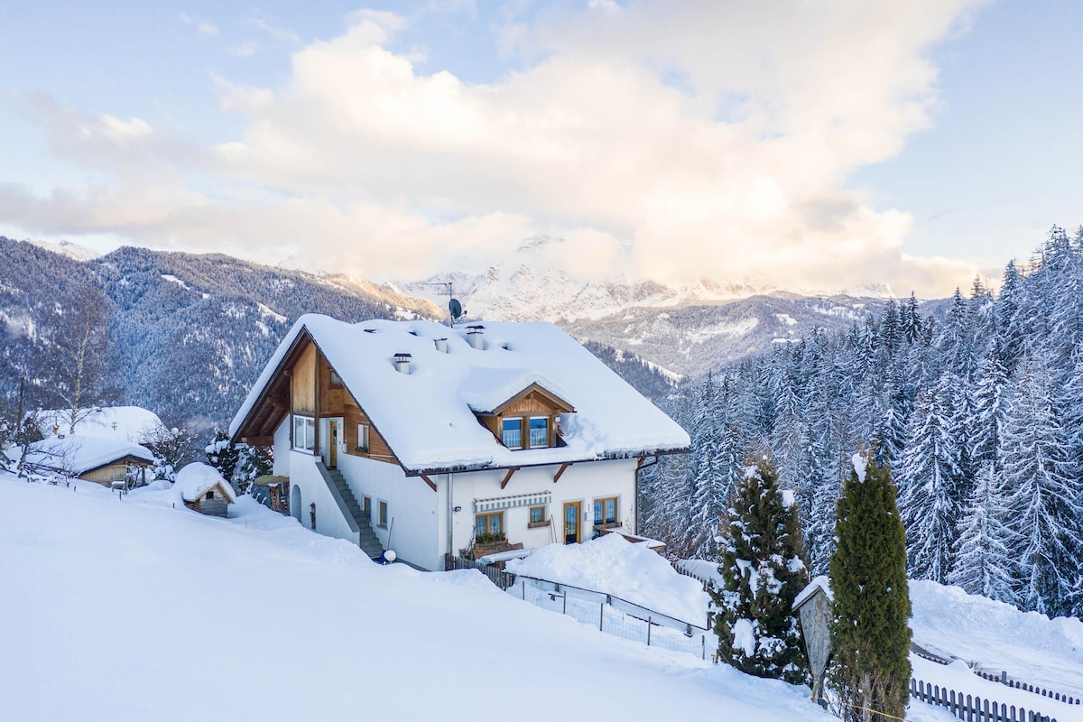 A charming house is nestled among a winter landscape, blanketed in fresh snow. Surrounding mountains tower in the background, with a mix of evergreen trees visible. Soft clouds drift by in a clear blue sky, creating a serene atmosphere.