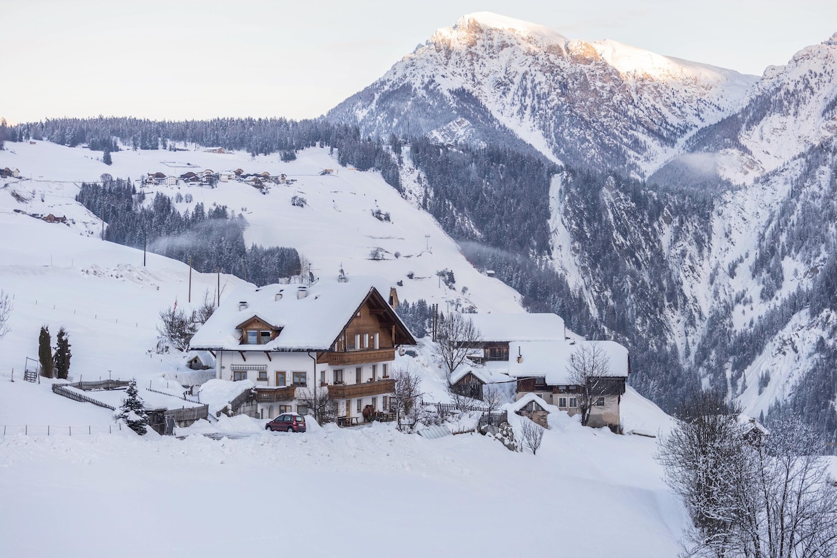 A scenic view captures snow-covered mountains rising in the background, with a traditional chalet nestled in the foreground. The tranquil landscape features trees adorned with snow and rolling hills, evoking a peaceful winter atmosphere.