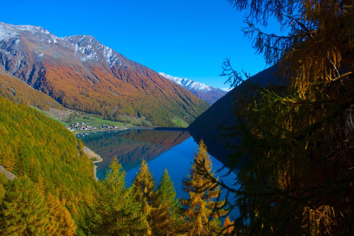 A panoramic view of a serene alpine landscape is captured, showcasing a clear lake bordered by vibrant autumn foliage. Majestic mountains rise in the background, with snow-dusted peaks contrasting against the blue sky. The tranquil waters reflect the surrounding trees and hills.