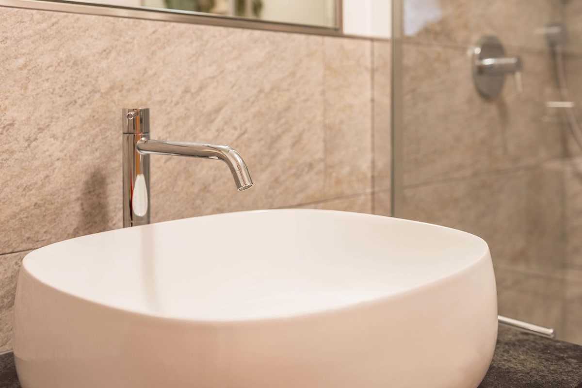 A modern bathroom sink is displayed, featuring a smooth, oval basin made of white material. A sleek, contemporary faucet extends over the sink, reflecting polished chrome finishes. The surrounding walls are covered in light-colored stone tiles, contributing to a clean and uncluttered aesthetic.