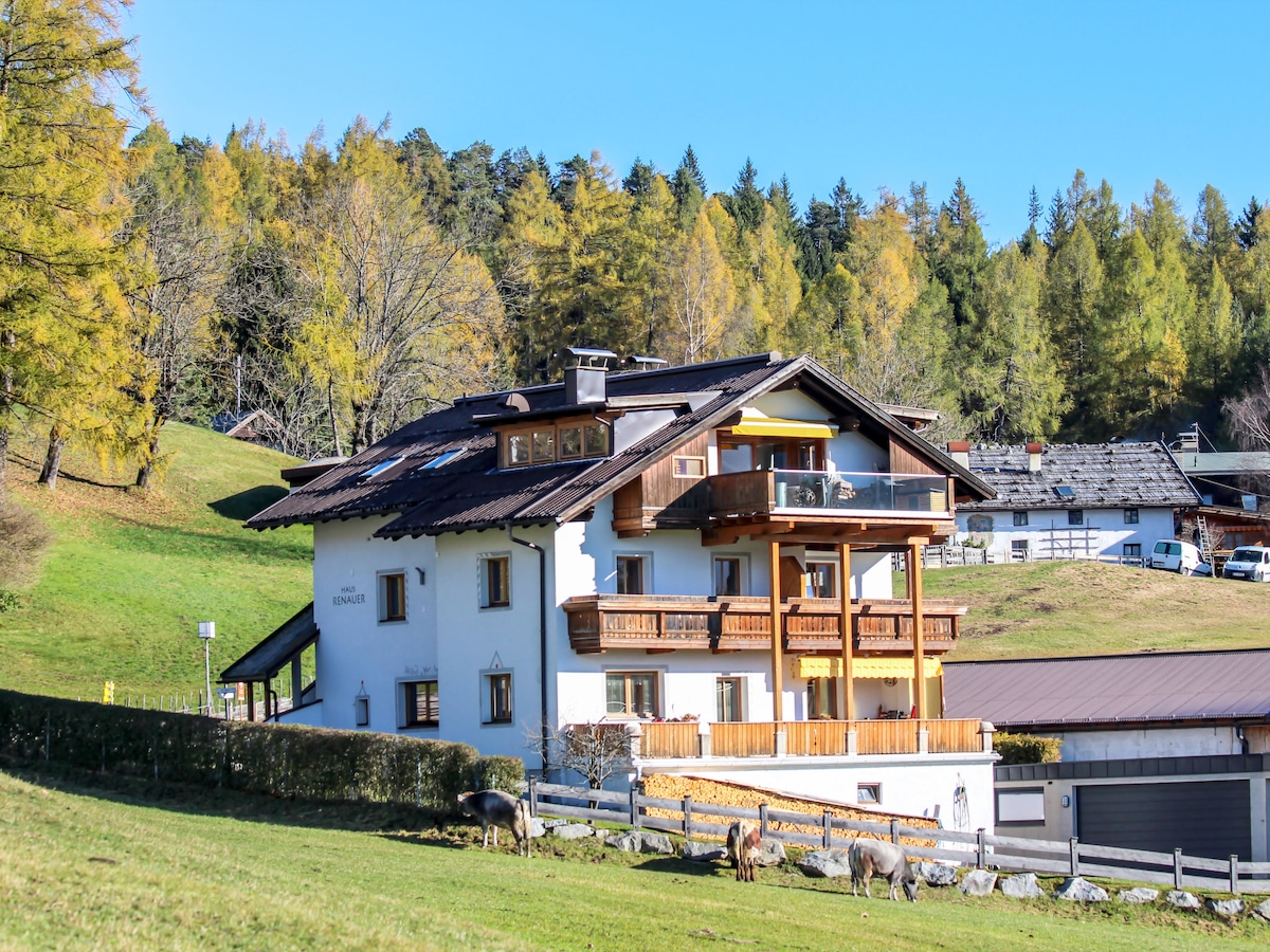 A spacious two-story apartment building is situated in a serene natural setting, surrounded by green meadows and trees in autumn hues. Balconies overlook the landscape, and several areas of the structure integrate wooden elements, blending traditional and modern designs.