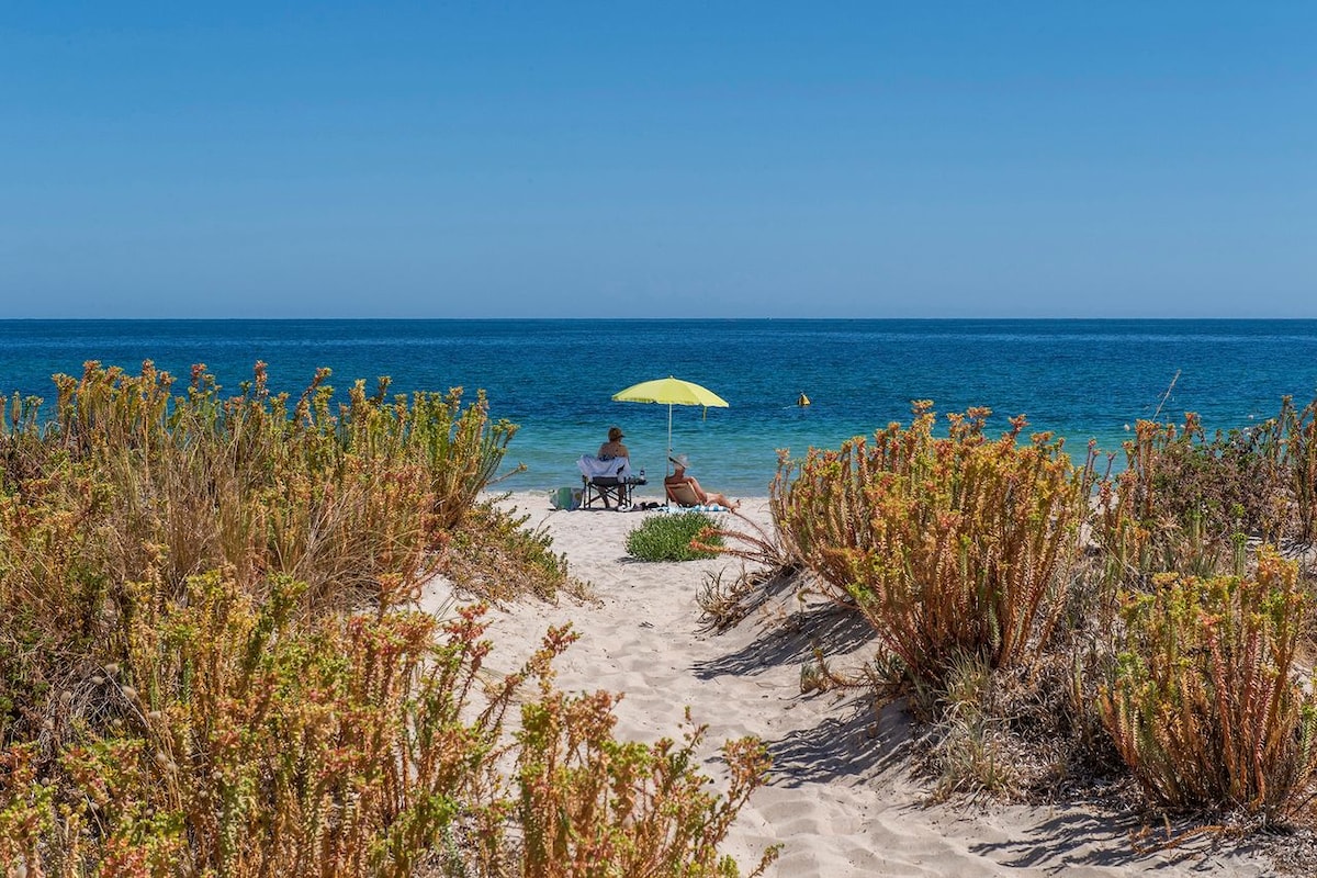 A peaceful beach scene is captured, featuring two individuals sitting under a bright yellow umbrella. Gentle waves lap at the shoreline, framed by sandy paths and lush coastal vegetation. The expansive blue sky stretches overhead, creating a serene atmosphere.