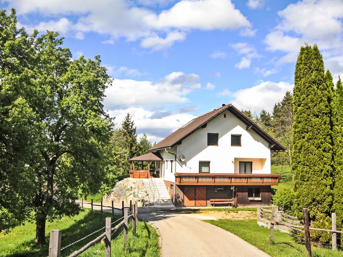 A detached three-room house is situated in a sunny position, surrounded by grassy meadows and fields. The exterior features a spacious entrance with stairs leading to a large balcony. Lush green trees frame the property, enhancing the countryside view.