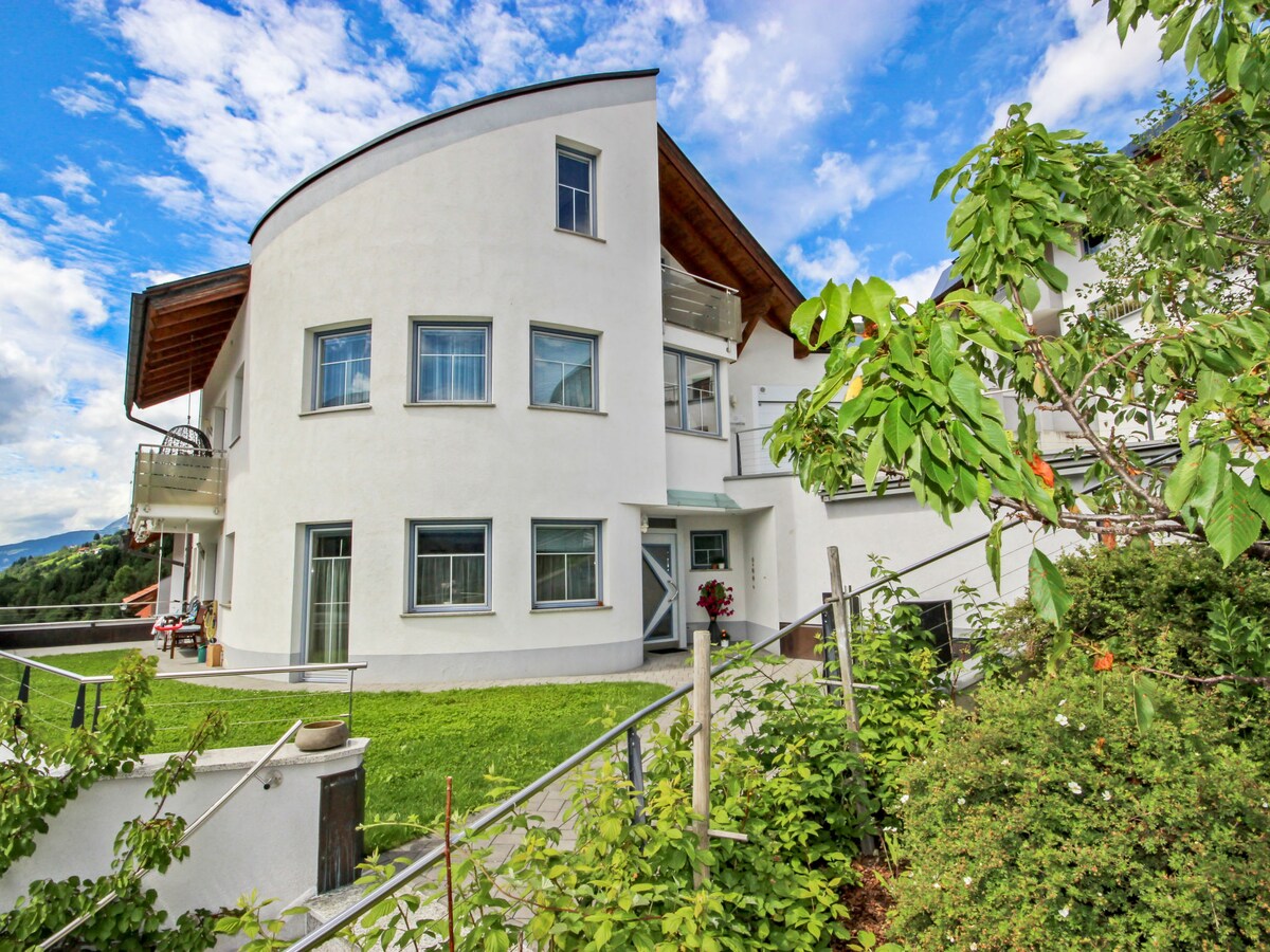 A modern three-story building is set against a vibrant sky, showcasing large windows and a curved façade. Lush greenery surrounds the entrance, complemented by well-maintained plants. A balcony is visible on the upper floor, offering an inviting outdoor space.