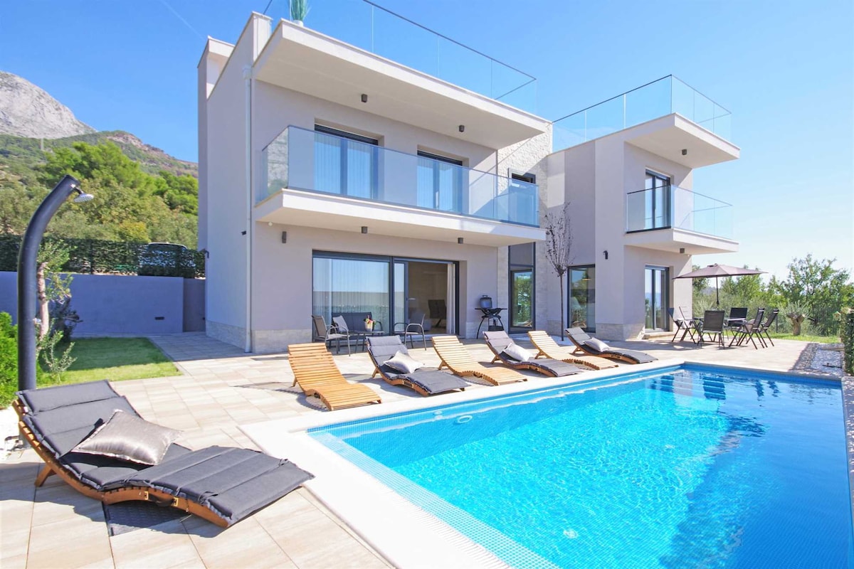 The exterior of the villa showcases a modern design with two levels and large glass doors. A trapezoidal swimming pool is surrounded by sunbeds and a sunbathing platform. Lush greenery and distant hills create a scenic backdrop, providing a relaxing atmosphere.