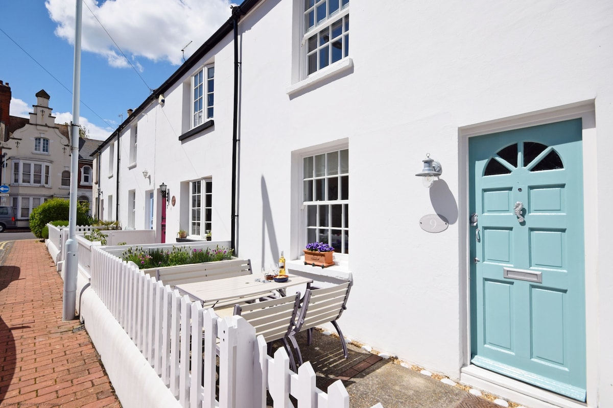 A charming cottage exterior is displayed with a white facade and a light blue front door. A quaint terrace area features a wooden table surrounded by chairs, complemented by flower boxes. The pathway is bordered by a white picket fence, enhancing the inviting appearance.