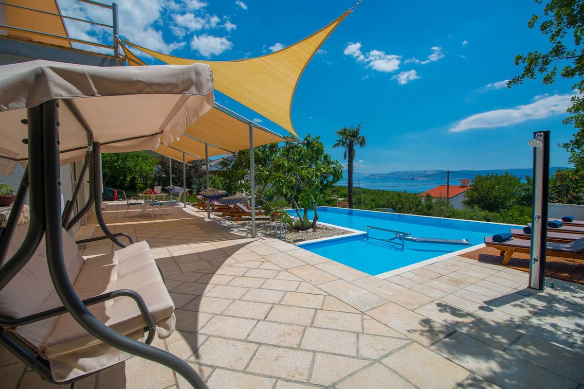 An outdoor space features a refreshing pool within a modern setting, accompanied by a shaded seating area and sun loungers. Lush greenery is visible, and a distant sea view enhances the tranquil environment. The sky is bright with scattered clouds, creating an inviting atmosphere.
