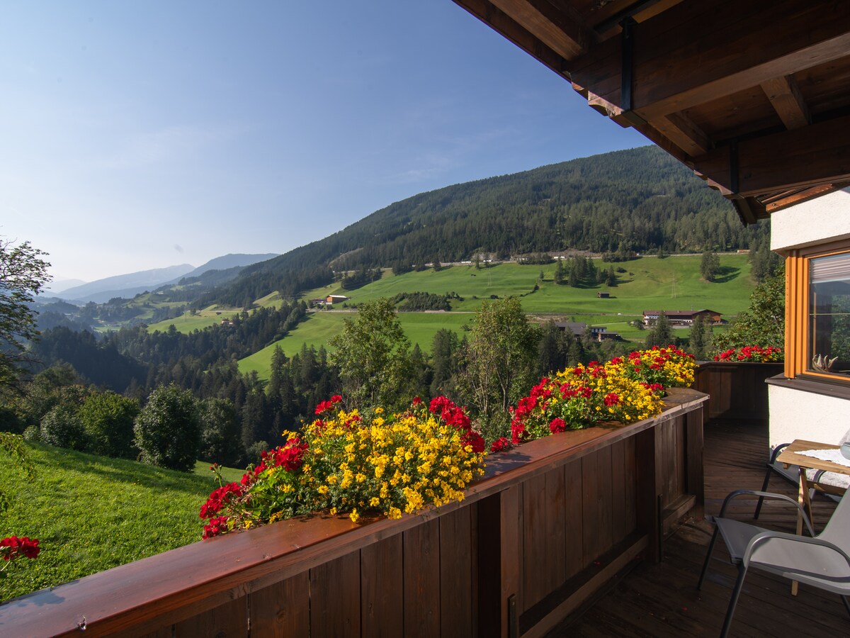 A wooden balcony adorned with vibrant flowers provides a panoramic view of the lush green landscape, rolling hills, and distant mountains. The tranquil scenery is bathed in natural light, highlighting the beauty of the surroundings.