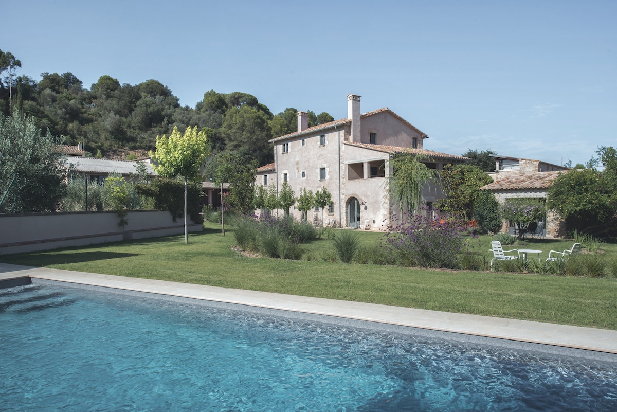 A spacious garden surrounds a charming stone house, with well-maintained grass and various trees. A refreshing pool is visible in the foreground, while seating areas complement the outdoor space, inviting relaxation and enjoyment of the natural surroundings.