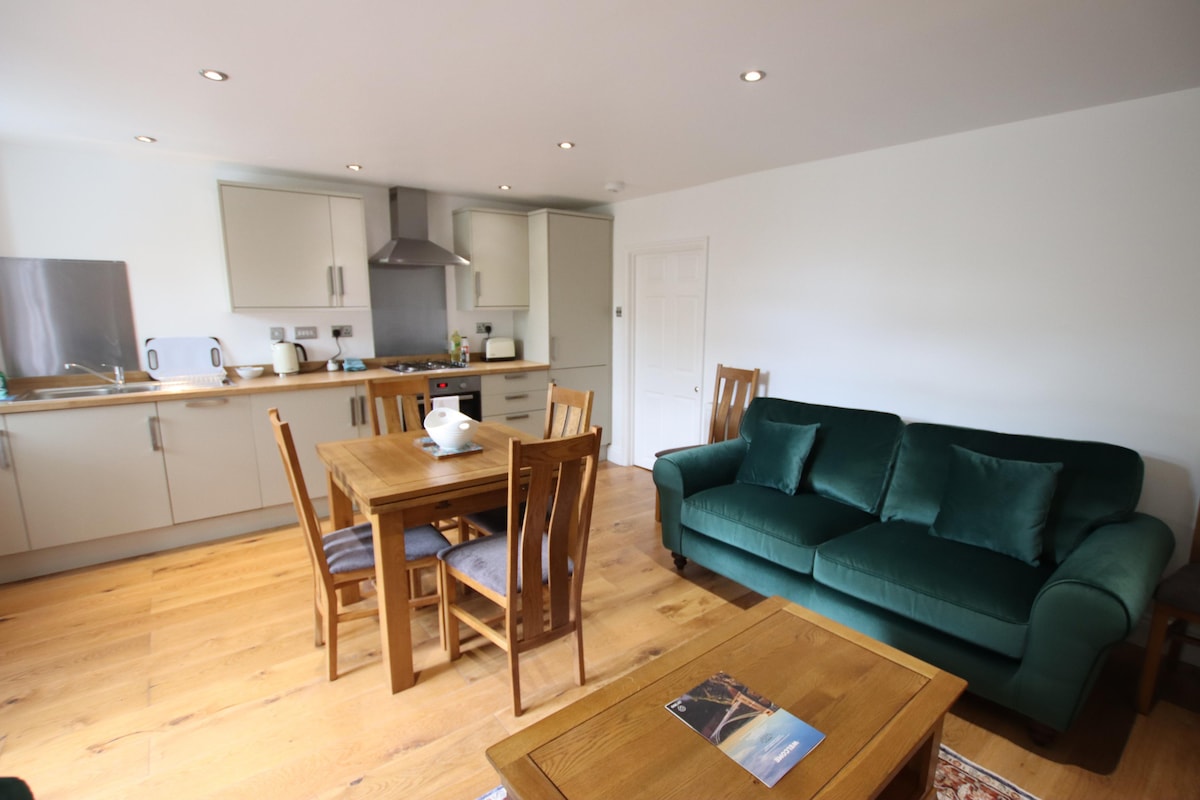 A bright open-plan living area is featured with a green sofa opposite a wooden dining table and chairs. A kitchen area is visible, equipped with modern appliances. Light hardwood floors create a warm ambiance, complemented by overhead recessed lighting.