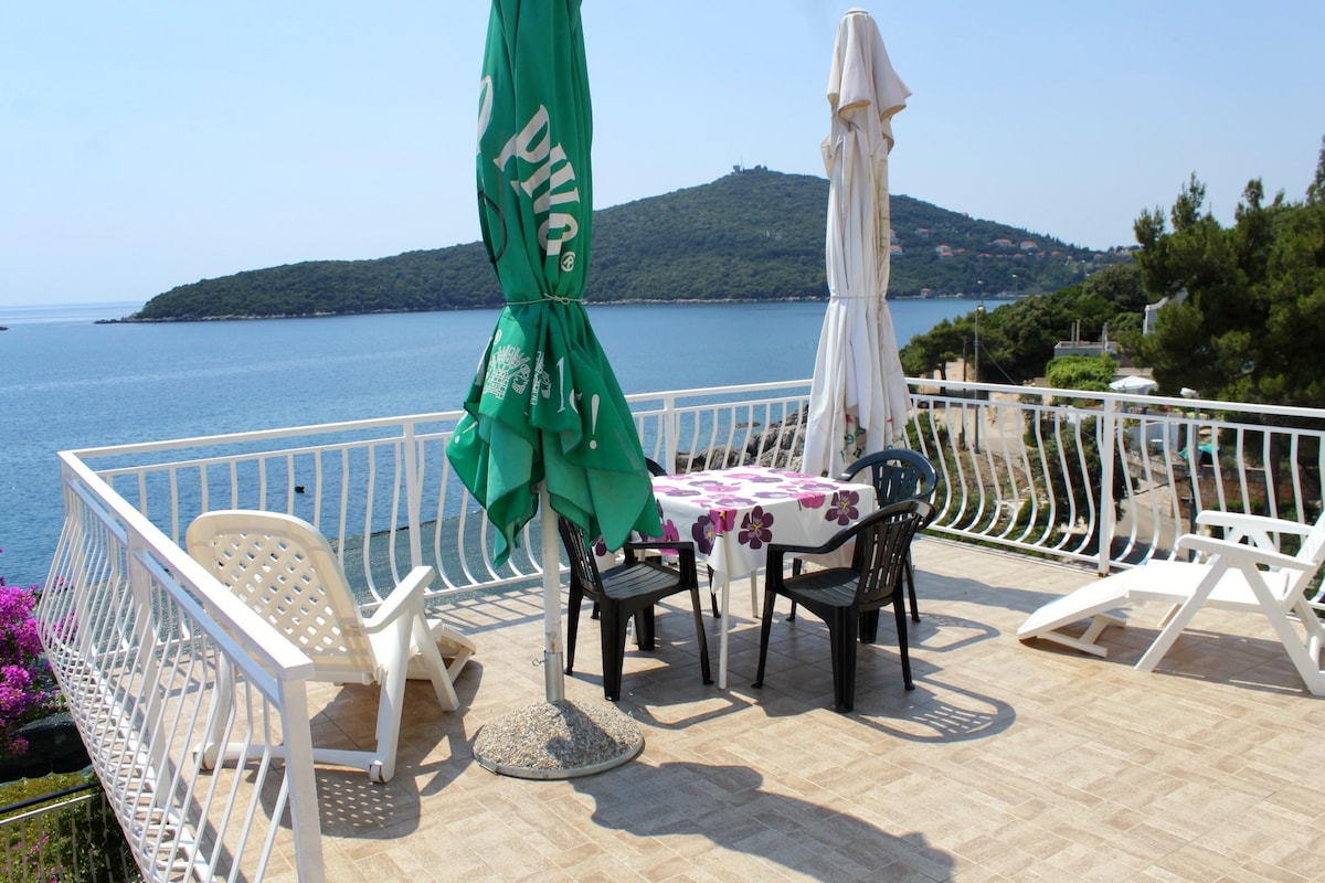 An outdoor terrace features a round table with chairs surrounded by sun loungers. Two umbrellas provide shade, and a scenic view of the sea and distant hills can be seen, creating an inviting spot for relaxation.