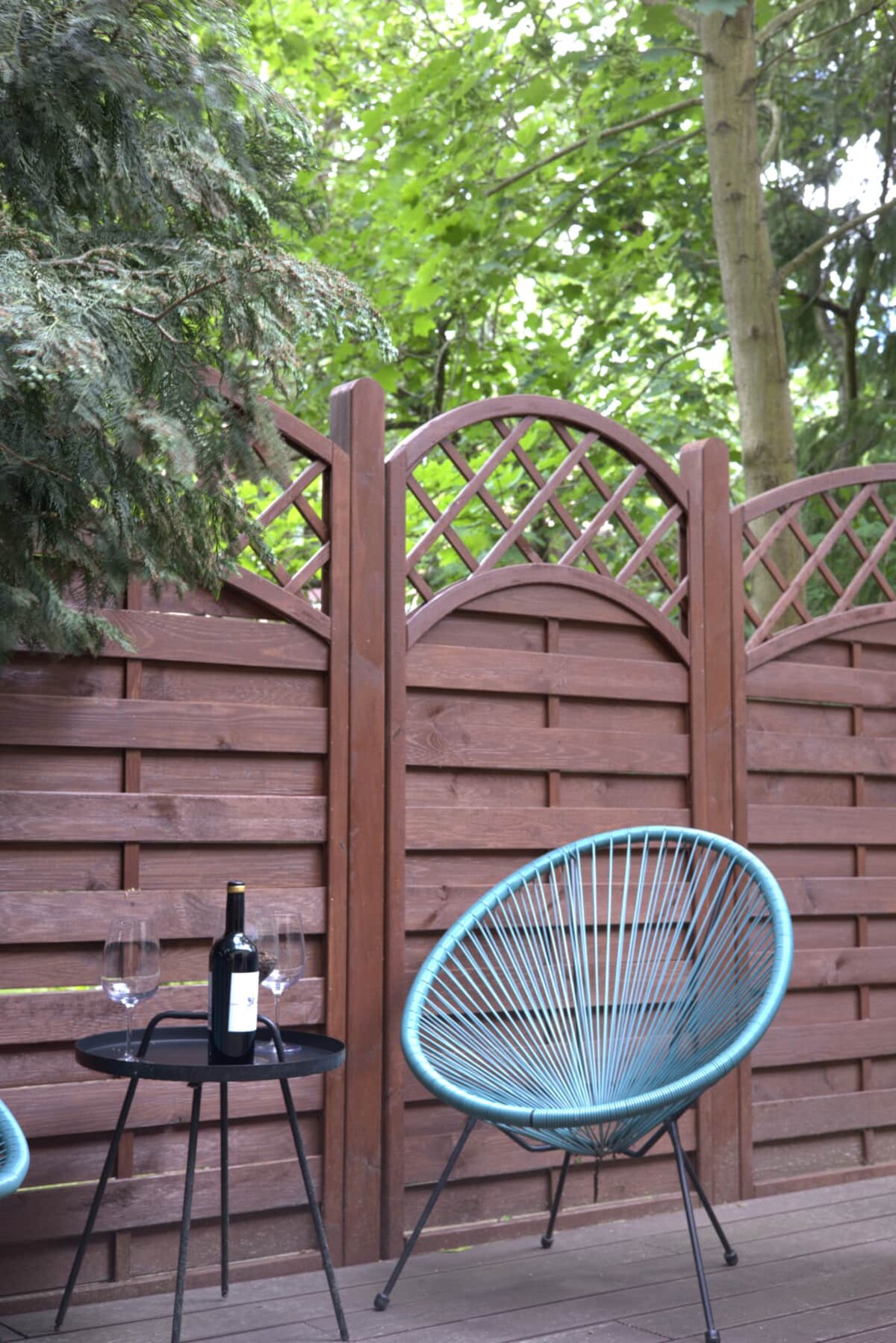 A corner of a garden terrace showcases two light blue chairs facing a small black table. A wine bottle and two glasses are positioned on the table. Lush greenery is visible in the background, and a wooden fence adds privacy.