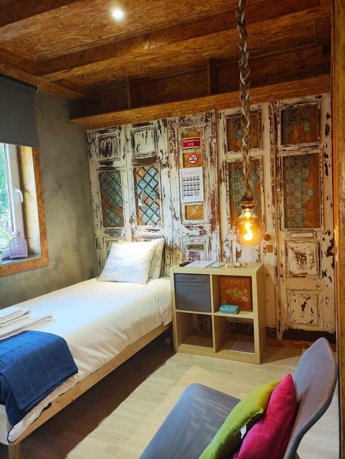 A cozy room features rustic wood paneling and an arrangement of decoratively aged doors. A single bed with white bedding is complemented by a soft blue throw. A small bedside table with storage sits next to the bed, and a bright pendant light hangs overhead.