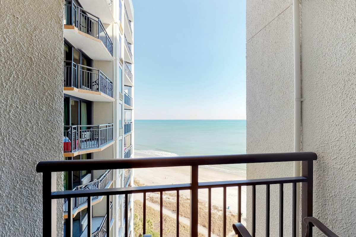 A view of the ocean and sandy beach is visible from the balcony, framed by the building's modern architecture. The tranquil blue water meets the horizon under a clear sky, creating a serene atmosphere.