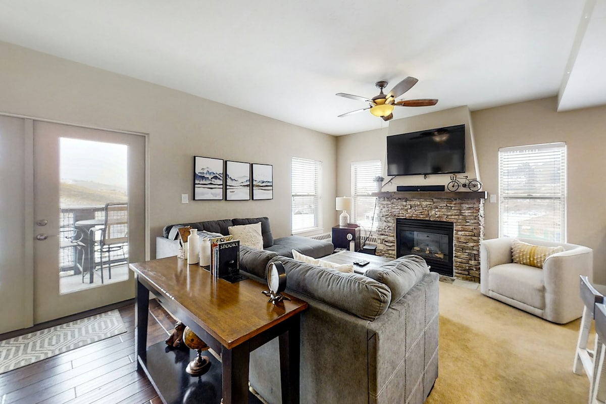 A living room is presented with a gas fireplace framed by natural stone, complemented by a ceiling fan. A spacious couch and lounge chair are arranged for relaxation. Large windows allow natural light to illuminate the room, with a sliding glass door offering access to outdoor views.