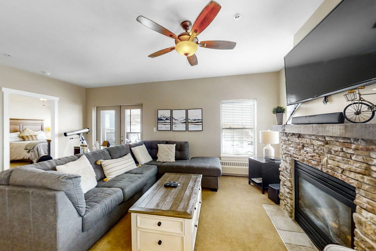 A spacious living area features a large gray sectional sofa and a rustic coffee table. A flat-screen TV is mounted above a stone fireplace. Natural light pours in through nearby windows, while artwork adorns the wall, complementing the neutral tones of the space.