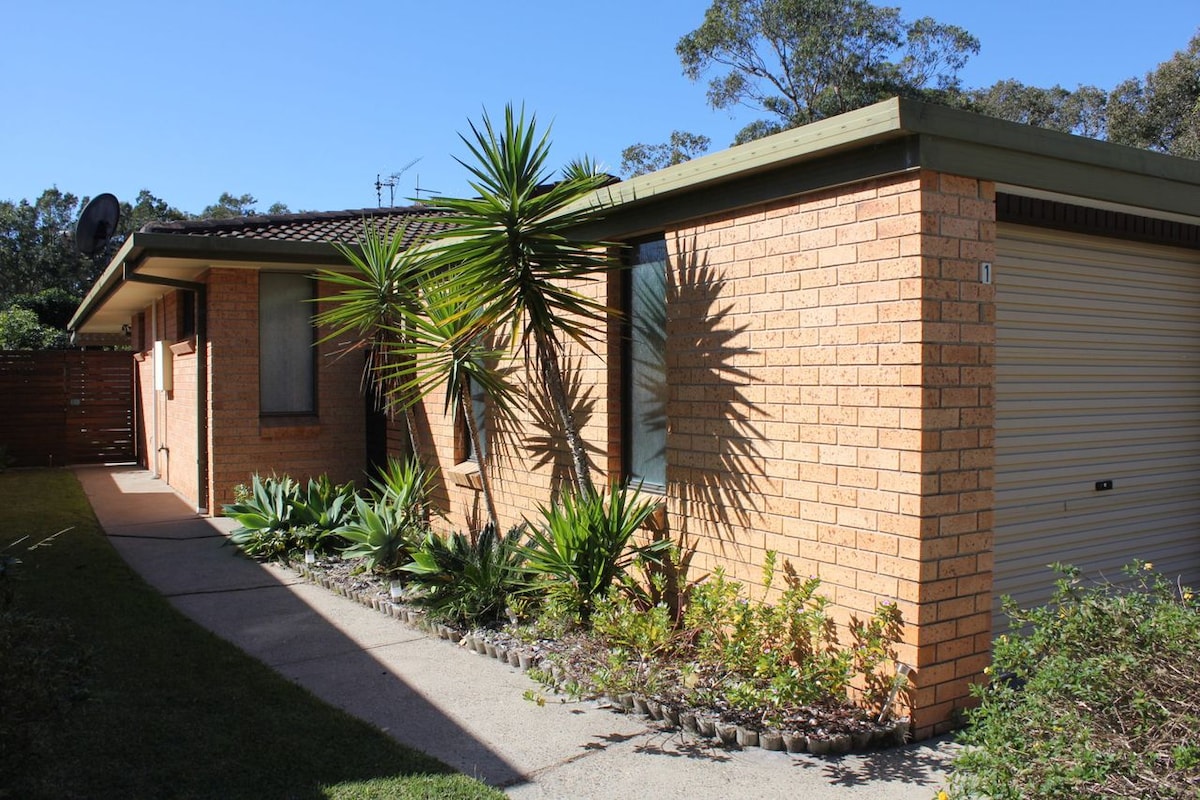 The exterior of the villa is displayed, featuring a brick façade and landscaped garden with various plants. A single-car garage with a roller door is visible, alongside a pathway leading to the entrance. Sunlight casts shadows of the foliage on the wall.