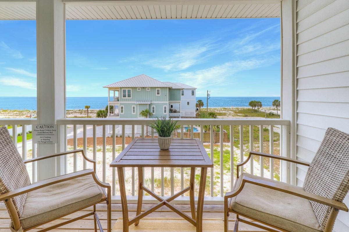 A private balcony is viewed, featuring a wooden table with two matching chairs. The ocean is visible in the background, complemented by a sandy landscape. A small plant adorns the table, enhancing the coastal ambiance.