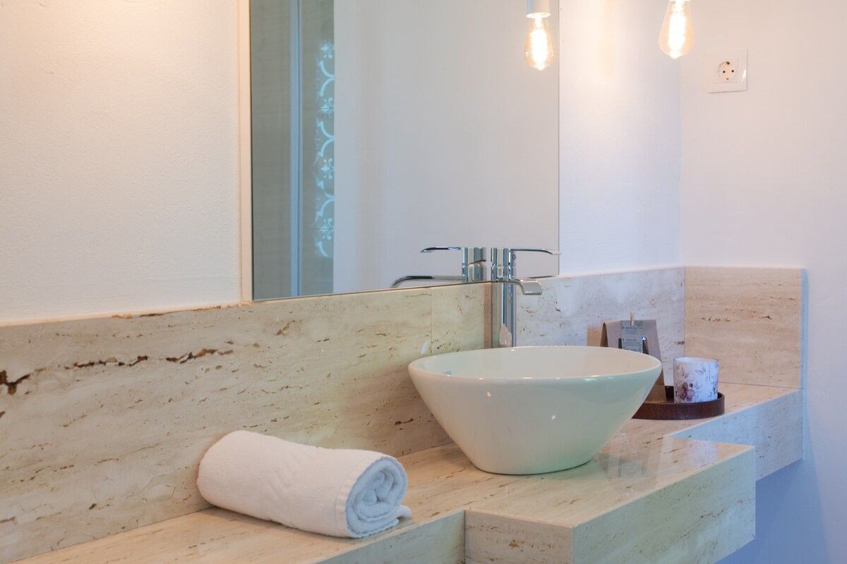 A modern bathroom features a stylish vessel sink placed on a sleek stone countertop. A hand towel is neatly rolled beside the sink. Soft lighting from hanging fixtures creates a serene ambiance, while a mirror reflects the minimalist design.