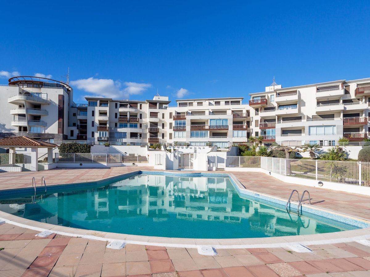 A shared swimming pool is set within a landscaped area, featuring a curved pool edge and surrounding sunbathing space. The complex buildings are visible in the background, under a clear blue sky. Poolside deck chairs are arranged for comfort and relaxation.