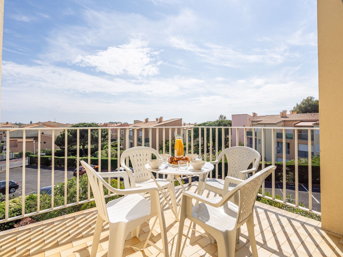 A small balcony with a table and four white chairs overlooks a garden area and other buildings. A breakfast setting is visible on the table, featuring drinks and pastries, with bright sunlight illuminating the space.