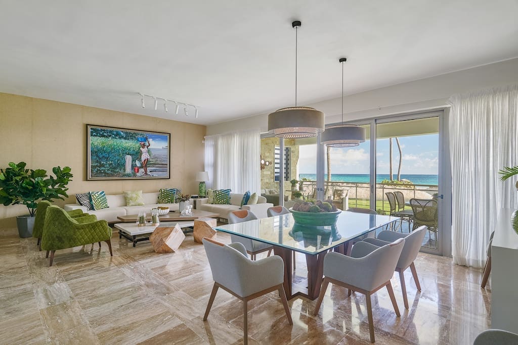 A spacious living area features a large dining table surrounded by stylish chairs. Comfortable seating is arranged around a center table, complemented by lush plants. Large sliding doors invite natural light and reveal ocean views, while a vibrant piece of artwork adorns the wall.