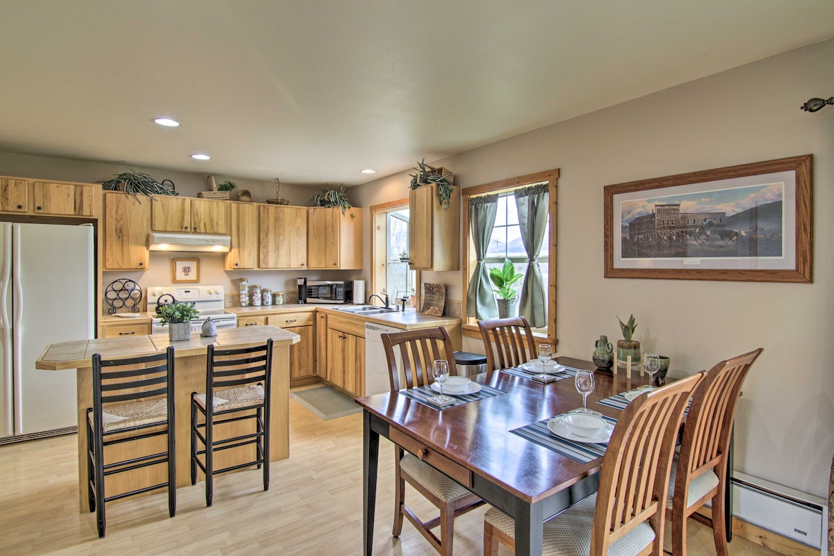 The kitchen and dining area feature wooden cabinetry, modern appliances, and ample counter space. A dining table is set for six, while four bar stools are positioned at the kitchen island. Natural light enters through the window, highlighting the functional layout.