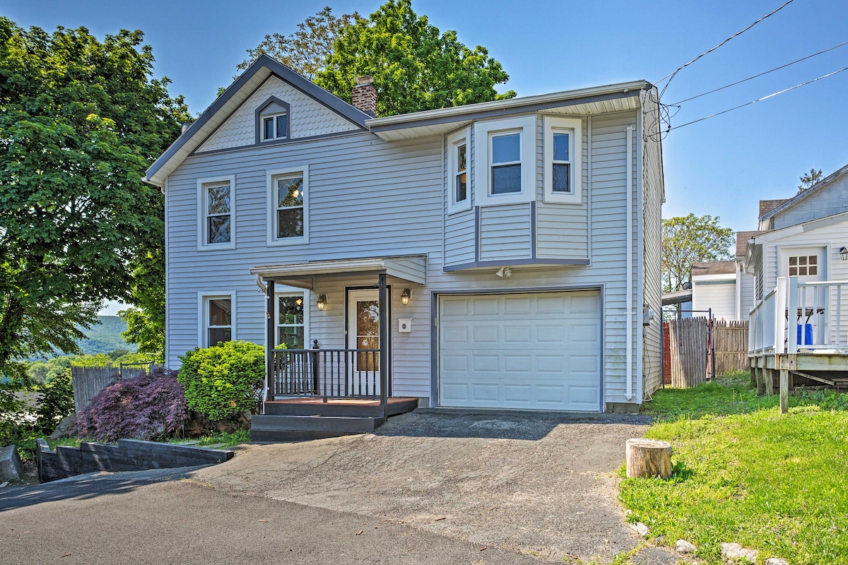 The exterior of the two-story home is shown with light grey siding and a welcoming front porch. A driveway leads to the garage, and lush green trees provide shade. The front yard is adorned with a flowering bush and a patch of grass.