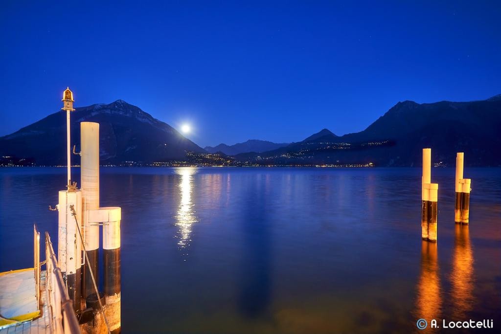 A serene lakeside scene is captured at night, featuring calm water reflecting the moonlight. Illuminated docks with tall white posts extend into the lake, framed by mountainous silhouettes. The surrounding hills are adorned with faint lights, suggesting distant settlements.