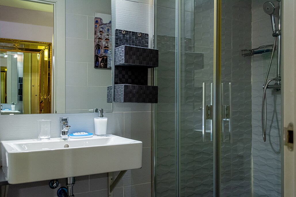 The bathroom features a modern design with a sleek sink supported by metal legs. A glass-enclosed shower is shown, along with decorative storage baskets mounted on the wall. Mirrors reflect light, enhancing the feeling of spaciousness.